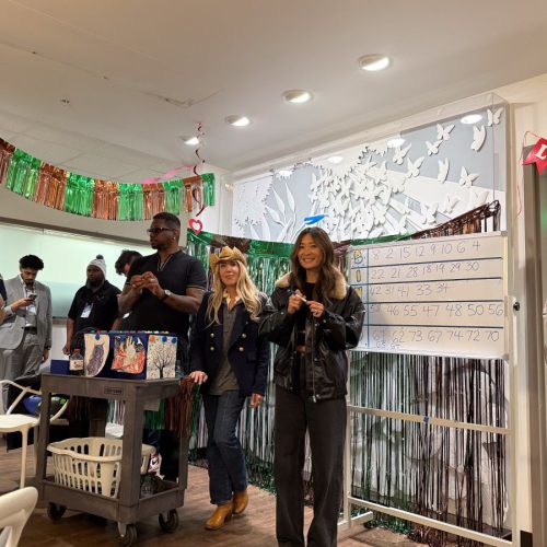 Photograph of a bingo game event with three people standing near a bingo cart holding cards and a microphone, while a large bingo board with numbered squares is displayed on a stand behind them. The setting includes festive decorations with green and orange streamers, white chairs, and a wall adorned with white paper butterflies and a sunburst design.