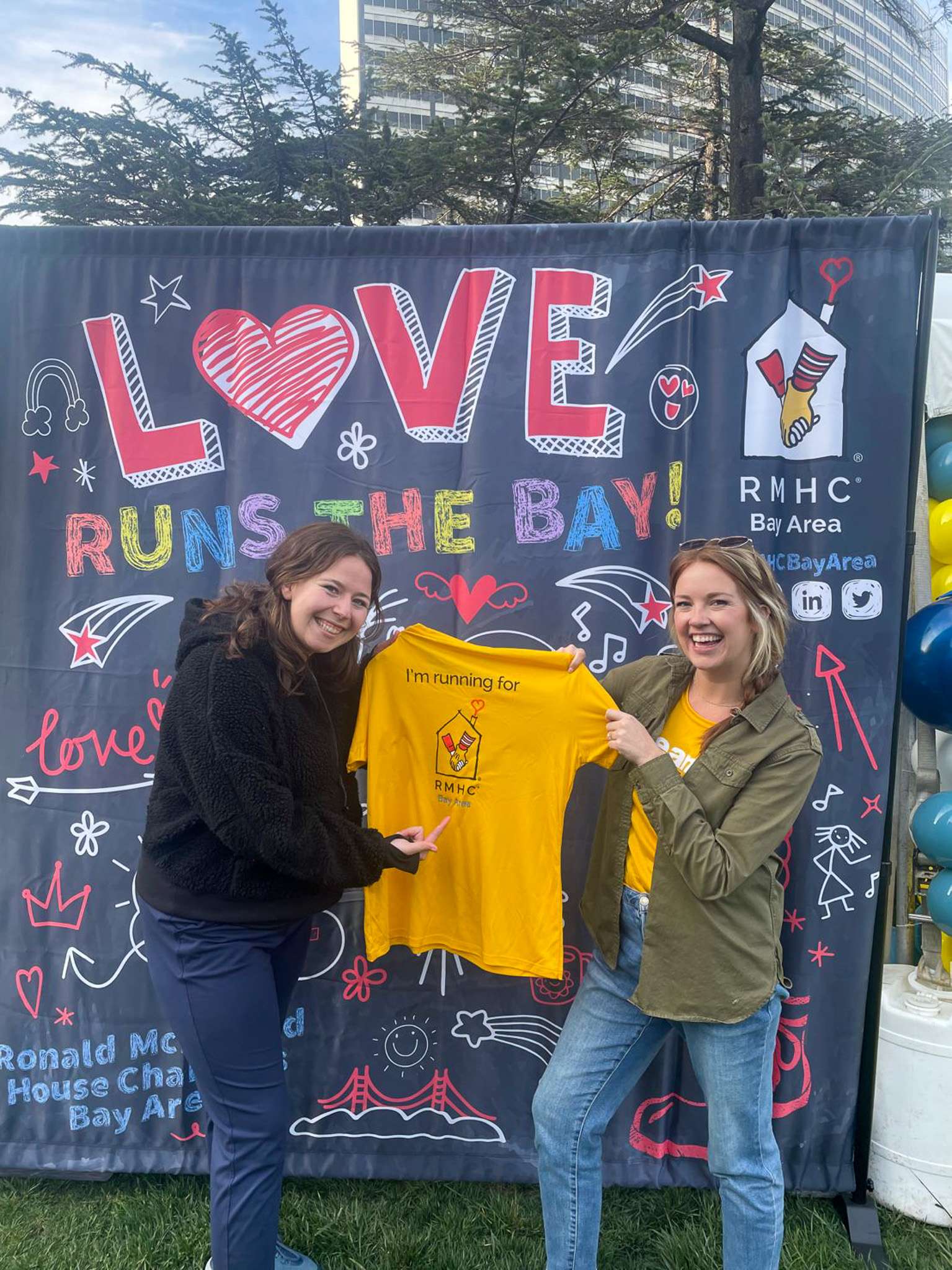 Two people holding a yellow Team RMHC shirt. There is a LOVE RMHC Bay Area backdrop