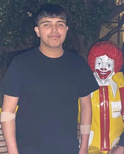 Teen with medical bandages on arms standing next to a Ronald McDonald statue at night.