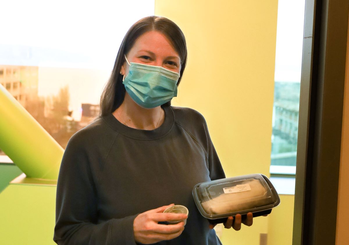 Feed the Love: RMHC Bay Area Guest Holding Packaged Meal Woman wearing a mask smiles while holding a packaged meal and sauce cup inside an RMHC facility.