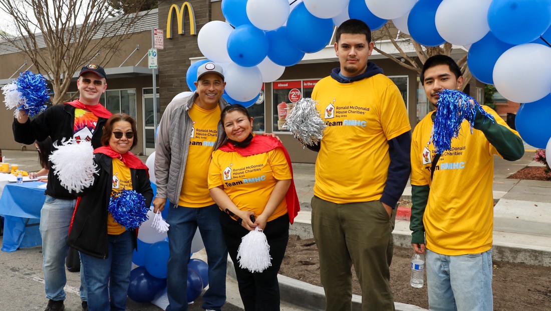 Group of RMHC volunteers in yellow shirts holding pom-poms and smiling under a blue and white balloon arch.