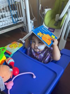 Smiling child with medical tape on their face holding a book over their head while sitting in a hospital chair surrounded by toys.