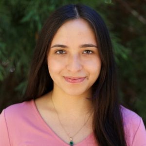 Agueda Gama Headshot. Woman with long dark hair wearing a pink top and green pendant necklace, smiling in front of greenery.