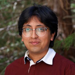 Headshot of Alan Gaspar Ruiz. Man wearing glasses smiling with a leafy green background.
