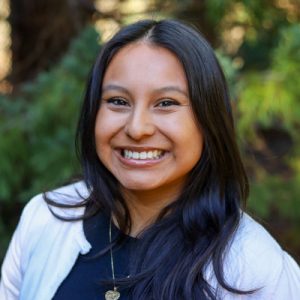 Andrea Carpano Headshot: Woman with smiling in front of greenery, wearing a black shirt with a white cardigan.
