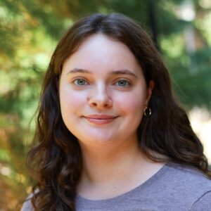 Ashley Mullin Headshot. Woman with wavy brown hair and gold hoop earrings smiling in front of a forested background.