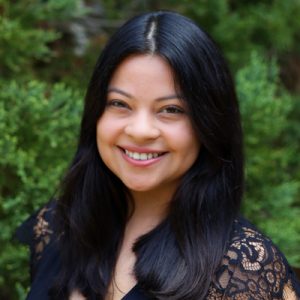 Smiling woman with long dark hair wearing a black lace top, standing outdoors in front of green foliage.