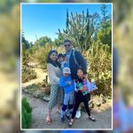 Family of five smiling in a cactus garden, with children holding toys and the parents standing close together in bright sunlight.