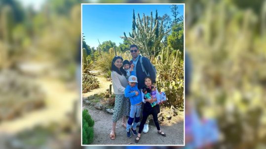 Family of five smiling in a cactus garden, with children holding toys and the parents standing close together in bright sunlight.