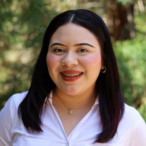 Smiling woman with straight dark hair, red lipstick, and a white blouse standing outdoors with trees in the background.