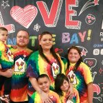 The Franco Family: Smiling family in rainbow tie-dye RMHC shirts posing together in front of Love Runs the Bay banner.