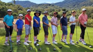 Group of young golfers wearing RMHC socks pose with golf clubs on a green course with mountain views.