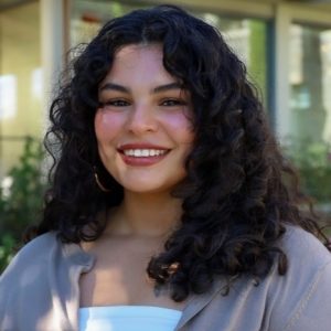 Headshot-Monse1-1 Monserrat Espinoza Headshot: Woman with curly hair and red lipstick smiling in front of greenery, wearing a white shirt with a brown cardigan.