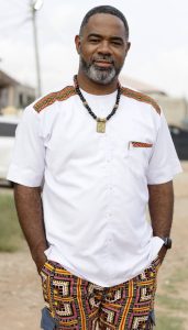 George Harris: Man wearing a white shirt with colorful trim, patterned shorts, and a black beaded necklace, standing outdoors.