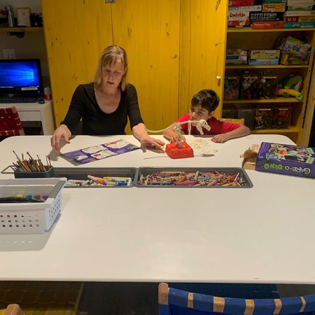 Jean, a Special Interest volunteer, and young boy sitting at a craft table with art supplies and a dinosaur model in Ronald McDonald House playroom.
