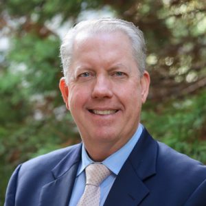 Jeff Rhode Headshot: Smiling man in a navy blazer and blue shirt standing against a green background.