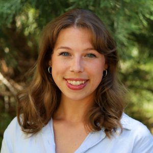 Lauren Keith Headshot. Woman with long light brown hair and blue eyes smiling outdoors while wearing a white shirt and chain necklace.