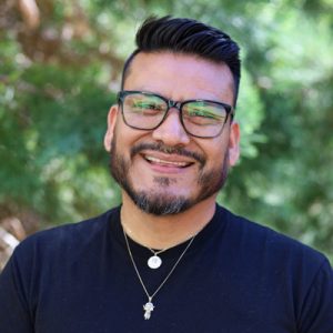 Headshot of Manuel. Man wearing glasses and a black shirt smiling. There are trees in the background.