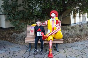 Smiling boy, Maxx, holding a sign and gift bag while sitting next to a Ronald McDonald statue on a bench outside RMHC.