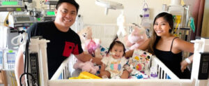 Parents smiling beside their toddler, Baby Adrianna, in a hospital crib surrounded by stuffed animals and medical equipment.