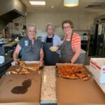 Three women wearing aprons smile while serving pizza slices in the Ronald McDonald House kitchen.
