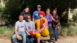 Izzy's family donating in honor of her. Group of smiling adults posing with a Ronald McDonald statue on a bench outside in a garden area.