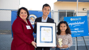 Chief Mission Officer, Bri Seoane with daughter, Izzy and Assembly Member Alex Lee. Three smiling individuals hold a certificate and trophy in front of a banner recognizing RMHC Bay Area as a 2021 Unsung Hero.