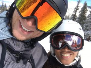 Smiling couple wearing ski helmets and goggles posing for a selfie on a snowy mountain.
