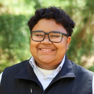 Rylee Quiambao Headshot. Person with short curly hair smiling in a brown polo shirt in front of green bushes.