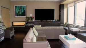 Modern waiting room with gray chairs, large TV, bookshelves, and natural light from tall windows.