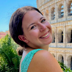 Keely Batmale. Woman in a green top smiles in front of the Colosseum in Rome on a sunny day.