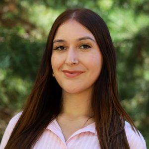 Stephanie Perez Headshot: Woman with long red hair and a nose piercing, wearing a brown top, standing in front of greenery.