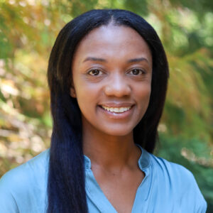 Tamara Garcia Headshot. Woman with long straight hair wearing a light blue top, smiling in front of leafy green trees.
