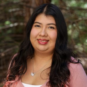 Yesenia Vazquez Headshot. Woman with long black hair smiles outdoors in front of a green leafy background.