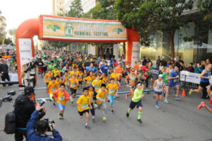 Large group of runners, including many children, starting the Oakland Running Festival under a colorful race banner.